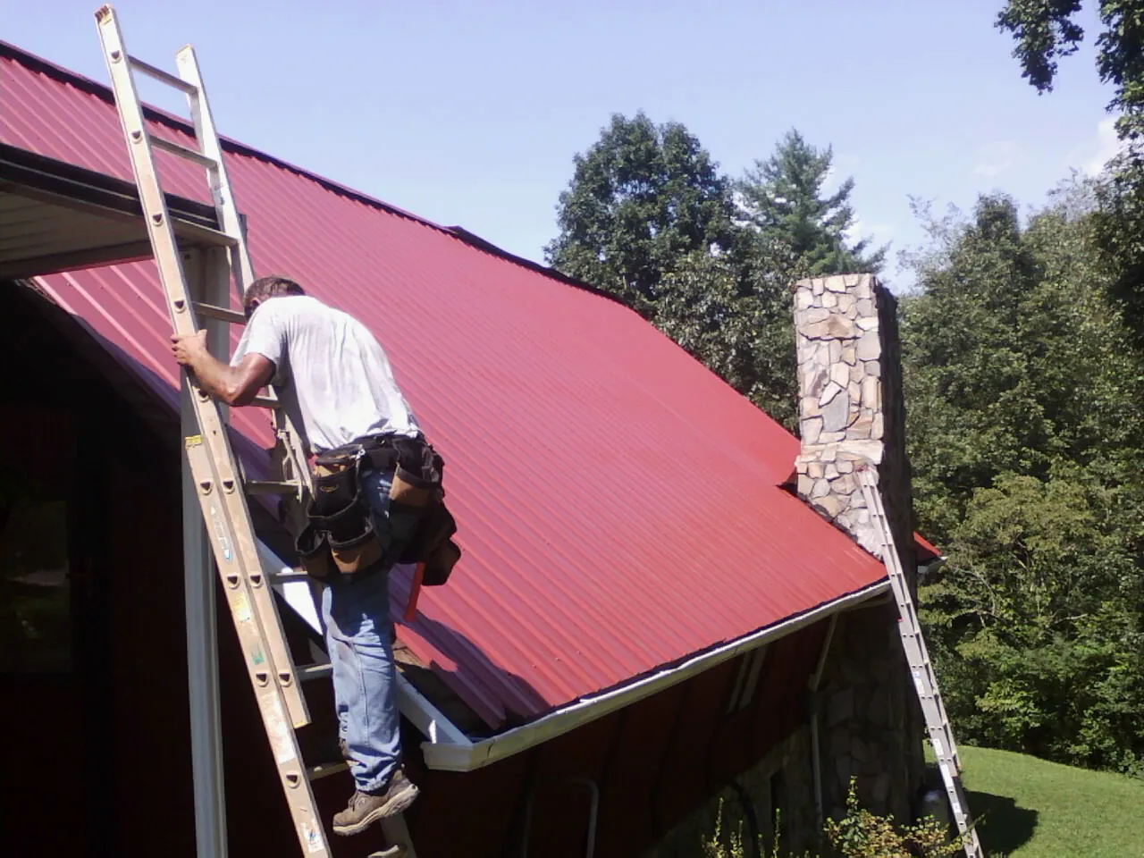 Roofer on a ladder beside a red metal roof and stone chimney — Soffit & Fascia Repair by Roofing Brothers in Calypso